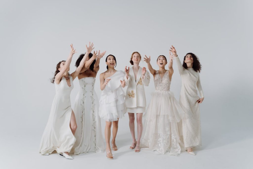 A diverse group of women joyfully tosses bouquets wearing stylish wedding dresses in a bright setting.