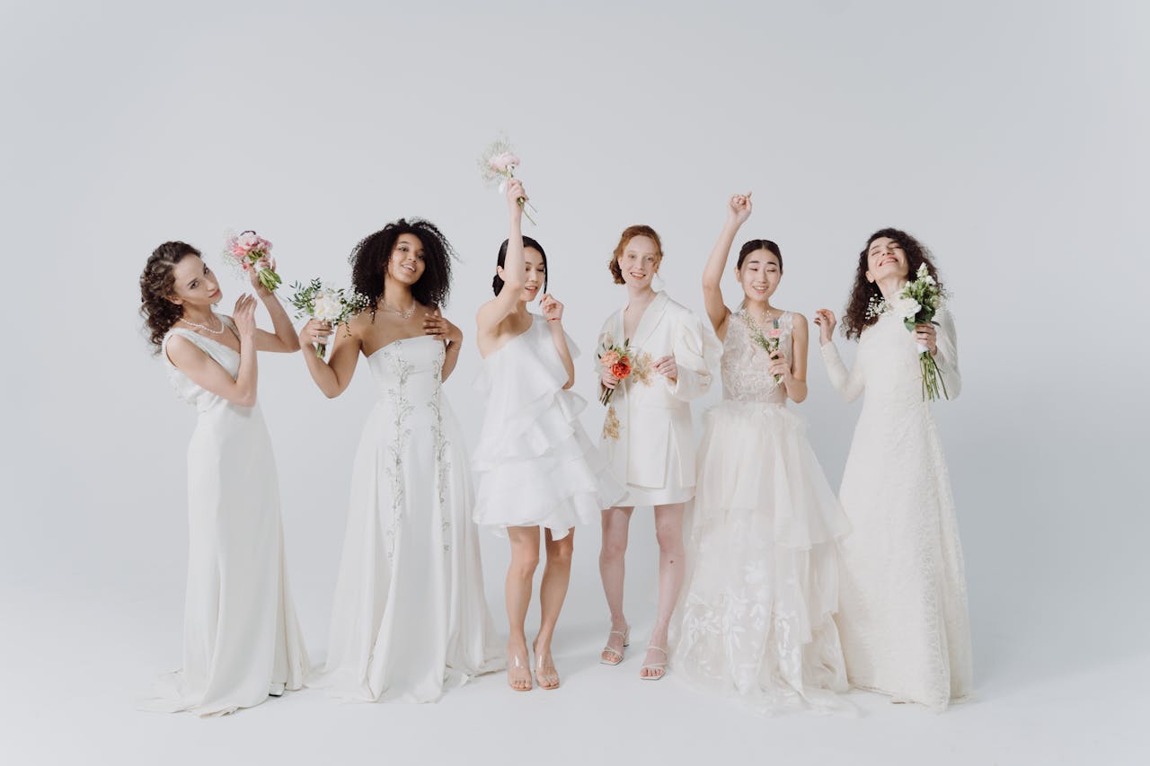 A group of diverse bridesmaids in white dresses holding bouquets, celebrating in a studio setting.